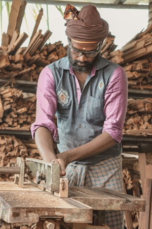 A man is working with a machine in a woodworking shop, wearing a turban and glasses. He is focused on the task at hand, surrounded by neatly stacked pieces of wood. The atmosphere appears to be industrious and traditional, reflecting a wood crafting process.