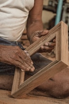 A person's hands are carefully sanding a wooden frame with sandpaper. The setting appears to be a workshop, with dust particles visible in the air and suggesting active woodworking.