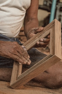 A person's hands are carefully sanding a wooden frame with sandpaper. The setting appears to be a workshop, with dust particles visible in the air and suggesting active woodworking.