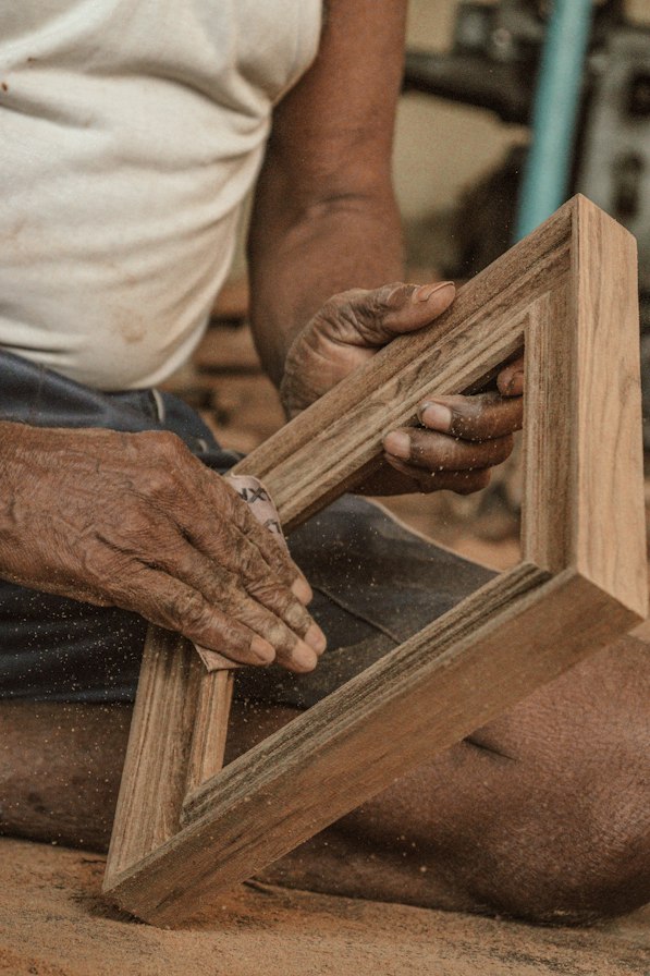 Close-up of hands sanding the edge of a wooden bookshelf panel with a sanding block in a bright workshop, smooth wood surface, dust particles in warm light