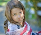 A young girl with long brown hair and green eyes looks towards the camera, partially wrapped in a flag-inspired shawl. The background is softly blurred with hints of green and yellow, suggesting an outdoor setting with sunlight filtering through.