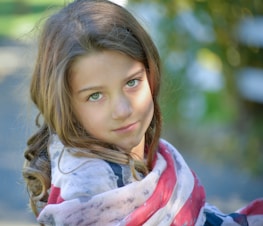 A young girl with long brown hair and green eyes looks towards the camera, partially wrapped in a flag-inspired shawl. The background is softly blurred with hints of green and yellow, suggesting an outdoor setting with sunlight filtering through.