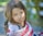 A young girl with long brown hair and green eyes looks towards the camera, partially wrapped in a flag-inspired shawl. The background is softly blurred with hints of green and yellow, suggesting an outdoor setting with sunlight filtering through.