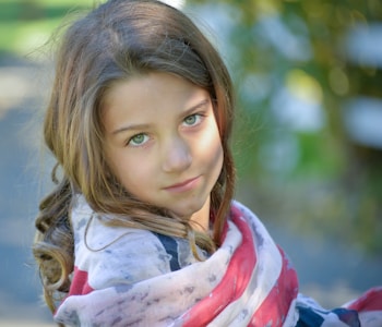 A young girl with long brown hair and green eyes looks towards the camera, partially wrapped in a flag-inspired shawl. The background is softly blurred with hints of green and yellow, suggesting an outdoor setting with sunlight filtering through.