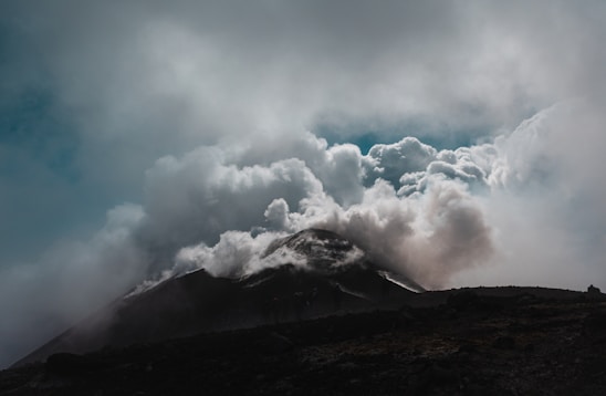 A large, dark volcanic mountain releases thick, billowing clouds of smoke and ash into a cloudy sky. The scene is dramatic, with sunlight partially breaking through the clouds, casting a shadow over the landscape.