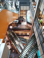 A multi-level shopping mall interior with several escalators intersecting at different angles. The escalators are surrounded by glass and metal railings, and modern spherical light fixtures hang from the ceiling. Shops and people can be seen on the various floors, and there is a spacious, open layout with visible seating areas.