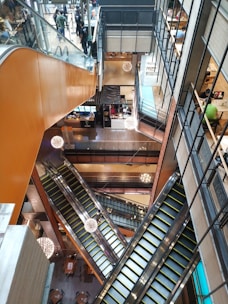 A multi-level shopping mall interior with several escalators intersecting at different angles. The escalators are surrounded by glass and metal railings, and modern spherical light fixtures hang from the ceiling. Shops and people can be seen on the various floors, and there is a spacious, open layout with visible seating areas.