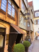 A charming, narrow street featuring a vintage-style hotel with exposed brickwork and wooden accents. The hotel sign is ornately decorated in black and gold, adding a classic touch. Bicycles are parked near the entrance, and a small vehicle is positioned on the side of the street. Large, neatly trimmed potted shrubs flank the entrance, providing a touch of greenery to the scene.