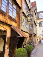 A charming, narrow street featuring a vintage-style hotel with exposed brickwork and wooden accents. The hotel sign is ornately decorated in black and gold, adding a classic touch. Bicycles are parked near the entrance, and a small vehicle is positioned on the side of the street. Large, neatly trimmed potted shrubs flank the entrance, providing a touch of greenery to the scene.