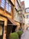 A charming, narrow street featuring a vintage-style hotel with exposed brickwork and wooden accents. The hotel sign is ornately decorated in black and gold, adding a classic touch. Bicycles are parked near the entrance, and a small vehicle is positioned on the side of the street. Large, neatly trimmed potted shrubs flank the entrance, providing a touch of greenery to the scene.