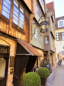 A charming, narrow street featuring a vintage-style hotel with exposed brickwork and wooden accents. The hotel sign is ornately decorated in black and gold, adding a classic touch. Bicycles are parked near the entrance, and a small vehicle is positioned on the side of the street. Large, neatly trimmed potted shrubs flank the entrance, providing a touch of greenery to the scene.