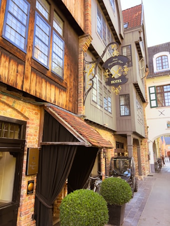 A charming, narrow street featuring a vintage-style hotel with exposed brickwork and wooden accents. The hotel sign is ornately decorated in black and gold, adding a classic touch. Bicycles are parked near the entrance, and a small vehicle is positioned on the side of the street. Large, neatly trimmed potted shrubs flank the entrance, providing a touch of greenery to the scene.