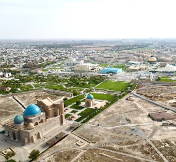 An aerial view of a cityscape with prominent historic architecture featuring blue domes. The foreground includes two large buildings with distinct domes, surrounded by a mix of green spaces and modern developments. The background showcases an expansive urban setting with numerous smaller buildings extending towards the horizon.