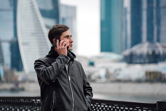 A man is standing outdoors in an urban setting, talking on his phone. He is wearing a black leather jacket and appears to be in a modern city area with tall buildings in the background.