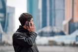 A person wearing a black leather jacket is holding a smartphone to their ear while standing outdoors. Skyscrapers with modern architecture are visible in the blurred background, suggesting an urban setting.
