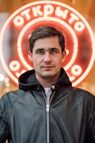 A person with short dark hair is wearing a black leather jacket with a zipped front. The background features a neon circular sign, glowing in red and white, with Cyrillic text.