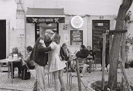 An outdoor seating area in front of a gelato shop features several people sitting at tables. A couple is sharing an affectionate embrace in the foreground. The shopfront is adorned with signage, and a menu board displays various gelato flavors and drinks. The mood is relaxed and social.