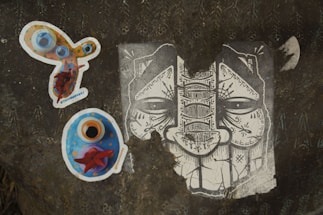 A dark textured surface displays three distinct pieces of artwork. On the left, there is a colorful, abstract sticker with circles and vibrant hues of orange, blue, and red. Below it, another sticker predominantly blue with orange and red accents appears to be some form of artistic depiction. On the right, a monochrome illustration of a face characterized by intricate patterns and symbols fills the surface, showcasing elaborate line work.