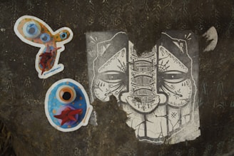 A dark textured surface displays three distinct pieces of artwork. On the left, there is a colorful, abstract sticker with circles and vibrant hues of orange, blue, and red. Below it, another sticker predominantly blue with orange and red accents appears to be some form of artistic depiction. On the right, a monochrome illustration of a face characterized by intricate patterns and symbols fills the surface, showcasing elaborate line work.