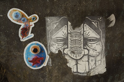 A dark textured surface displays three distinct pieces of artwork. On the left, there is a colorful, abstract sticker with circles and vibrant hues of orange, blue, and red. Below it, another sticker predominantly blue with orange and red accents appears to be some form of artistic depiction. On the right, a monochrome illustration of a face characterized by intricate patterns and symbols fills the surface, showcasing elaborate line work.