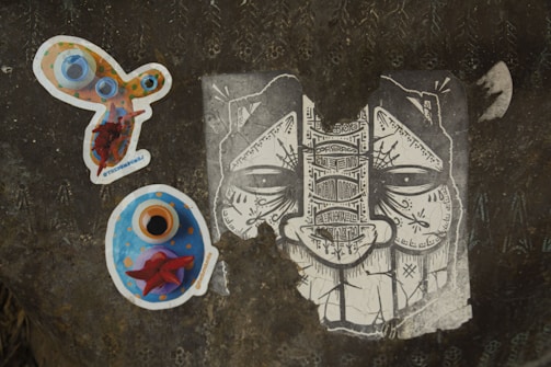 A dark textured surface displays three distinct pieces of artwork. On the left, there is a colorful, abstract sticker with circles and vibrant hues of orange, blue, and red. Below it, another sticker predominantly blue with orange and red accents appears to be some form of artistic depiction. On the right, a monochrome illustration of a face characterized by intricate patterns and symbols fills the surface, showcasing elaborate line work.