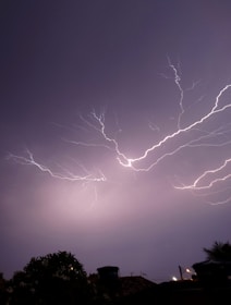 Bright lightning bolts streak across a dark night sky, illuminating the scene with intense light. Silhouetted trees and rooftops are visible at the bottom, grounding the dramatic display above.