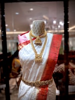A mannequin displays a traditional South Asian saree featuring a white fabric with golden polka dots and a bold red border. The saree is adorned with intricate gold jewelry, including a necklace and a waist belt. The background appears to be a richly decorated interior with ambient lighting.