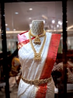 A mannequin displays a traditional South Asian saree featuring a white fabric with golden polka dots and a bold red border. The saree is adorned with intricate gold jewelry, including a necklace and a waist belt. The background appears to be a richly decorated interior with ambient lighting.