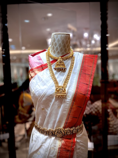 A mannequin displays a traditional South Asian saree featuring a white fabric with golden polka dots and a bold red border. The saree is adorned with intricate gold jewelry, including a necklace and a waist belt. The background appears to be a richly decorated interior with ambient lighting.