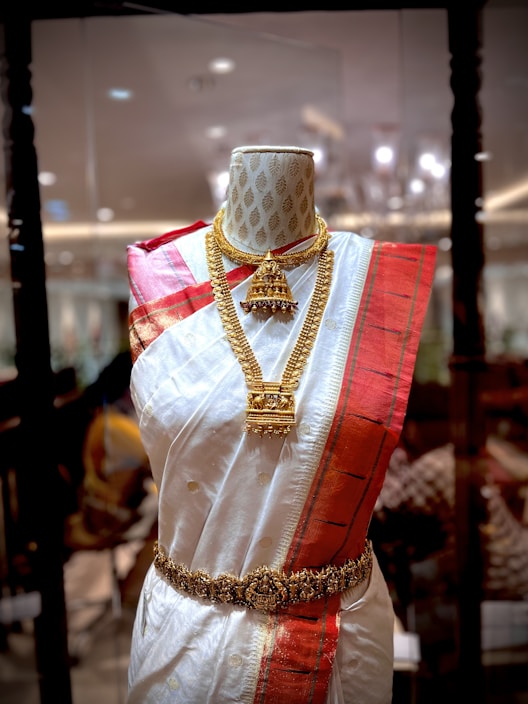 A mannequin displays a traditional South Asian saree featuring a white fabric with golden polka dots and a bold red border. The saree is adorned with intricate gold jewelry, including a necklace and a waist belt. The background appears to be a richly decorated interior with ambient lighting.