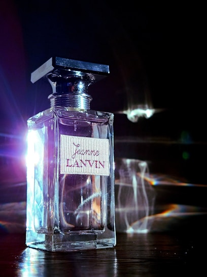 A clear glass perfume bottle labeled 'Jeanne Lanvin' with a decorative cap sits on a reflective surface. Bright light and lens flare effects create a dramatic illumination, highlighting the bottle's defined edges and casting colorful reflections.