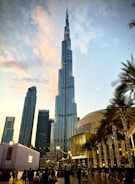A modern cityscape features a towering skyscraper at the center, surrounded by other high-rise buildings. The sky has a blend of blue and orange hues as the sun sets. Palm trees and festive lights adorn the foreground, where people are walking and gathering.