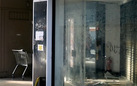 An abandoned storefront features graffiti-covered windows and walls. Inside, the area is dimly lit with reflections and scattered debris. A shopping cart is positioned on the left side, adding to the sense of neglect. Flyers are pasted on the surface beside the cart, and a fire extinguisher is visible inside.