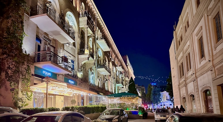 A vibrant nighttime city scene with elegant, illuminated building facades featuring ornate balconies and windows. The street is lined with parked cars and people walking, while overhead, strings of lights create a festive atmosphere. The signage of a restaurant or store named 'D'Olivo' is visible, adding to the lively urban setting.