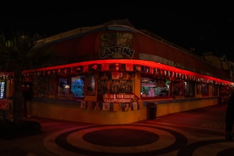 A nighttime scene of a colorful cantina with bright red LED lights adorning the perimeter of the roof. Various flags and banners are hung around the building, and there are promotional signs including one for an 'All You Can Eat Taco Tuesday'. The interior appears moderately lit with visible tables and some decor. A palm tree is visible to the left.