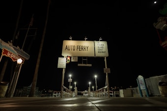 An entrance to an auto ferry is depicted. Prominent features include a large illuminated sign reading 'AUTO FERRY' and 'TO BALBOA ISLAND COAST HIGHWAY'. There are traffic lights and various signage visible, along with streetlights illuminating the area. The surroundings are dark, suggesting it is nighttime. To the left, a signboard reading 'U-DRIVE' is partially visible. The scene leads towards a dock area with barriers and railings.