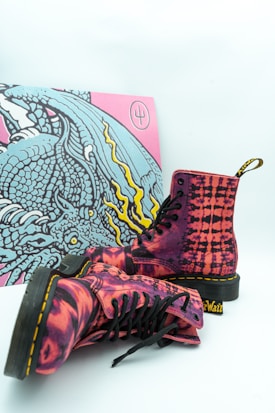 A pair of vibrant, colorful boots with a striking pink and black pattern is placed in front of a vivid artwork depicting a highly detailed, stylized dragon with turquoise, yellow, and pink hues. The boots have black laces and soles, with the brand's name visible on the heel loop.