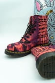 A pair of vibrant, patterned boots with a distinctive red, pink, and black tie-dye design. The boots feature black laces and sturdy black soles with yellow stitching. A background with a colorful, abstract design is visible behind the boots.