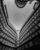 A dramatic architectural perspective showcasing the inner courtyard of a building with tall brick walls and numerous windows. The view points upwards, creating a sense of height and depth as the building seems to curve slightly inward, forming a partial enclosure of the sky.