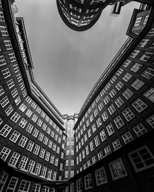 A dramatic architectural perspective showcasing the inner courtyard of a building with tall brick walls and numerous windows. The view points upwards, creating a sense of height and depth as the building seems to curve slightly inward, forming a partial enclosure of the sky.