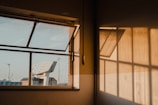 An open window in a dimly lit room lets in sunlight, creating warm shadows on the wall. Visible through the window is a modern building with angular design and wind turbines in the distance. The scene conveys a mix of natural and industrial elements under a golden sunlight.