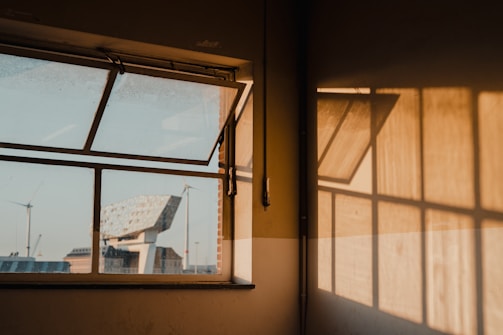 An open window in a dimly lit room lets in sunlight, creating warm shadows on the wall. Visible through the window is a modern building with angular design and wind turbines in the distance. The scene conveys a mix of natural and industrial elements under a golden sunlight.