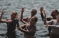 A group of people standing in water are engaged in a dance or meditative activity, with their eyes closed and hands raised in expressive gestures.