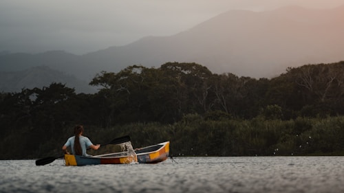 A person paddles a colorful kayak on a calm lake with dense green forest bordering the water. In the background, misty mountains create a serene and atmospheric backdrop under a soft, diffused light.