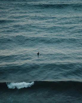 An individual with a surfboard is alone in the expansive ocean, surrounded by gentle, rippling waves. The sea extends to the horizon, creating a sense of vastness and solitude.