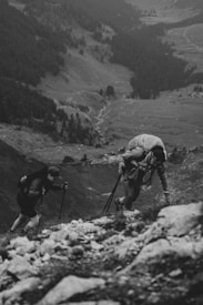 Two hikers are climbing a rocky and steep mountain path while using trekking poles. They carry large backpacks, and the background reveals a vast valley with a mix of trees and open fields, creating a dramatic and expansive natural setting.