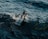 A scuba diver is partially submerged in a vast expanse of ocean water. The diver is equipped with a full scuba diving gear, including oxygen tanks and a black diving suit. Water splashes around the diver as waves move across the surface.