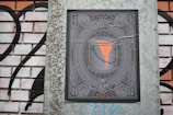 A detailed illustration of a paint bucket with orange paint dripping from it is centered within an ornate frame, reminiscent of currency notes. The design is displayed on a brick wall with black and white graffiti in the background.