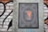 A detailed illustration of a paint bucket with orange paint dripping from it is centered within an ornate frame, reminiscent of currency notes. The design is displayed on a brick wall with black and white graffiti in the background.