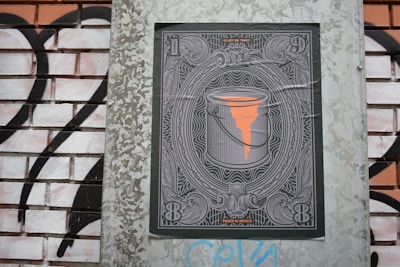 A detailed illustration of a paint bucket with orange paint dripping from it is centered within an ornate frame, reminiscent of currency notes. The design is displayed on a brick wall with black and white graffiti in the background.